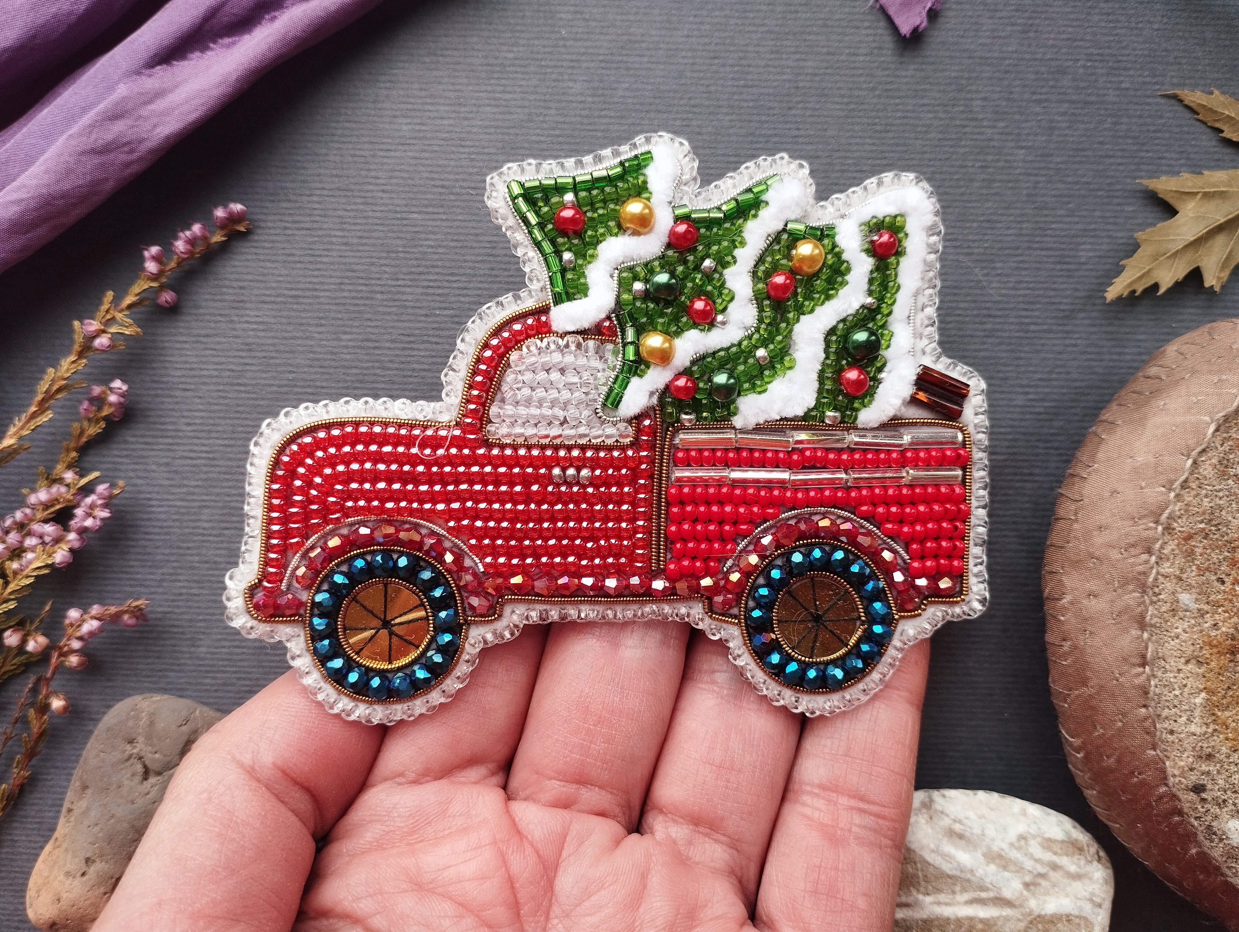 A hand is holding a red and white embroidered Christmas tree in a truck, surrounded by various objects such as leaves, flowers, and rocks.
